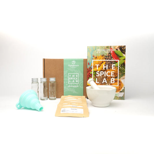 Collection of the content of the spice lab kit including a box, spices, funnel, bottles, a mortar and pestle, and a book titled 'The Spice Lab' on a white background.