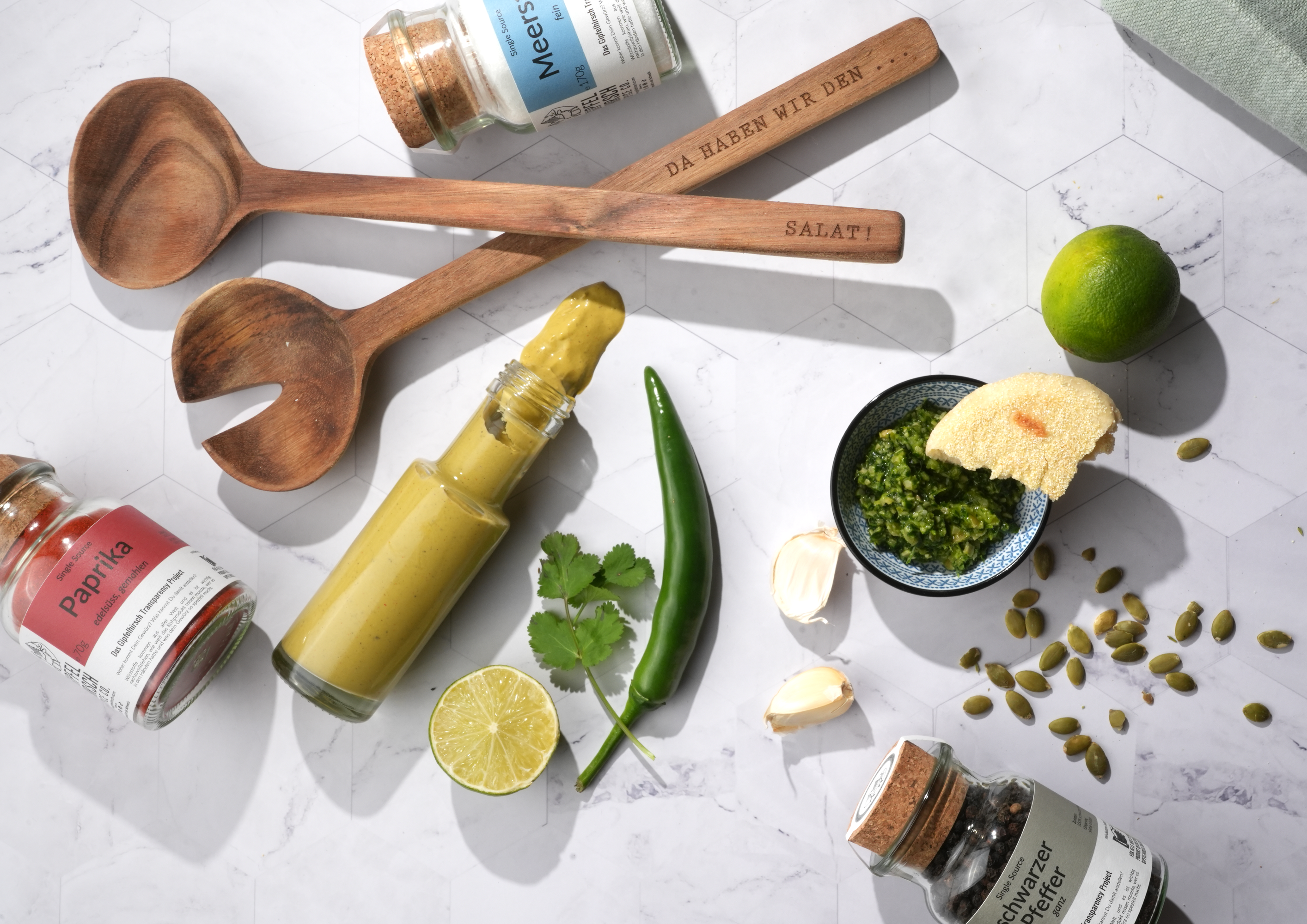 A Flatlay image with Gipfelhirsch Spice Products arranged on a white background. A green chili, lime and coriander leaves showcase the raw ingredients that were used to make a ipping sauce.